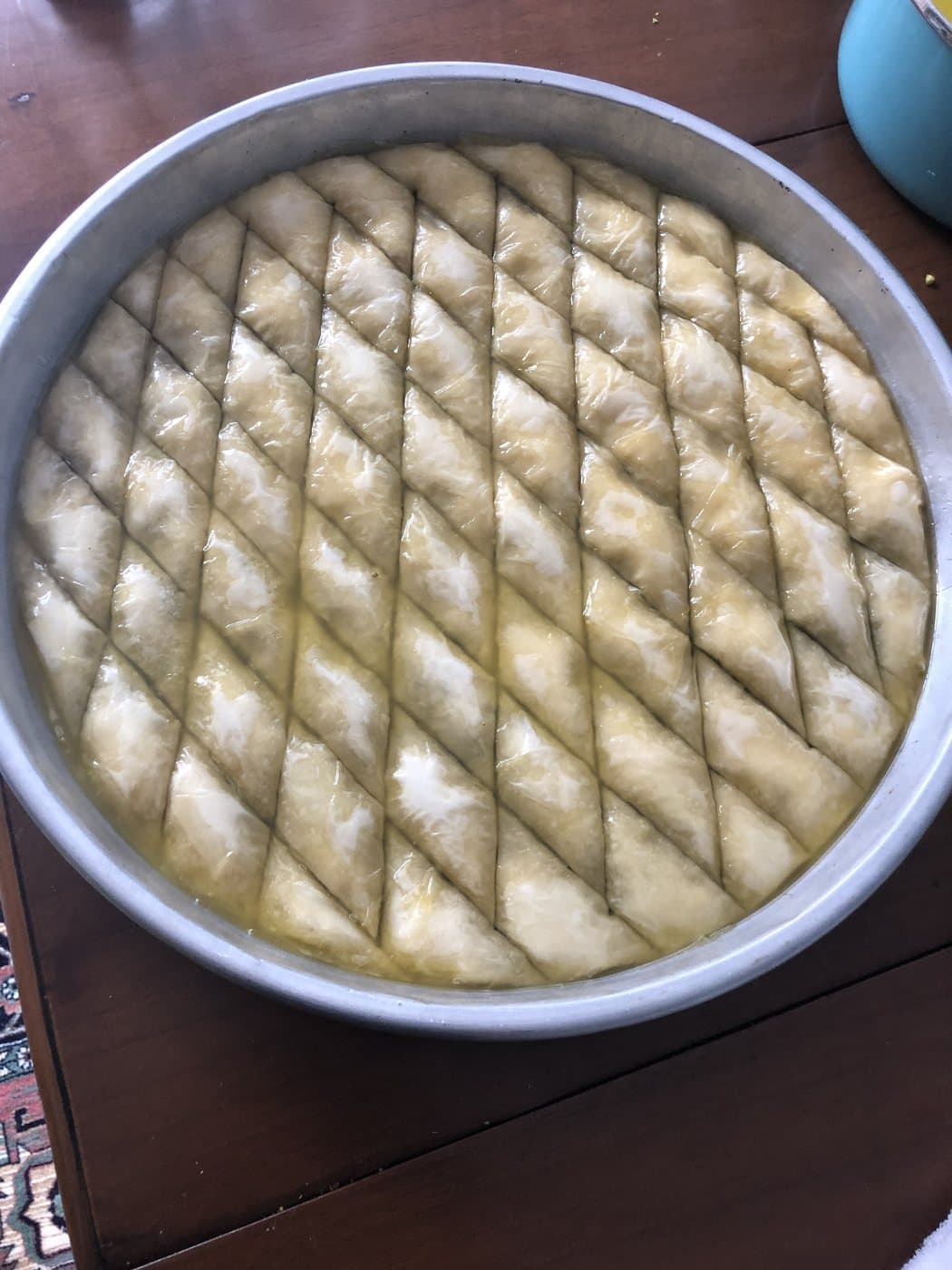 A round tray of unbaked baklava scored into diamonds and pooled with butter, on a wooden table