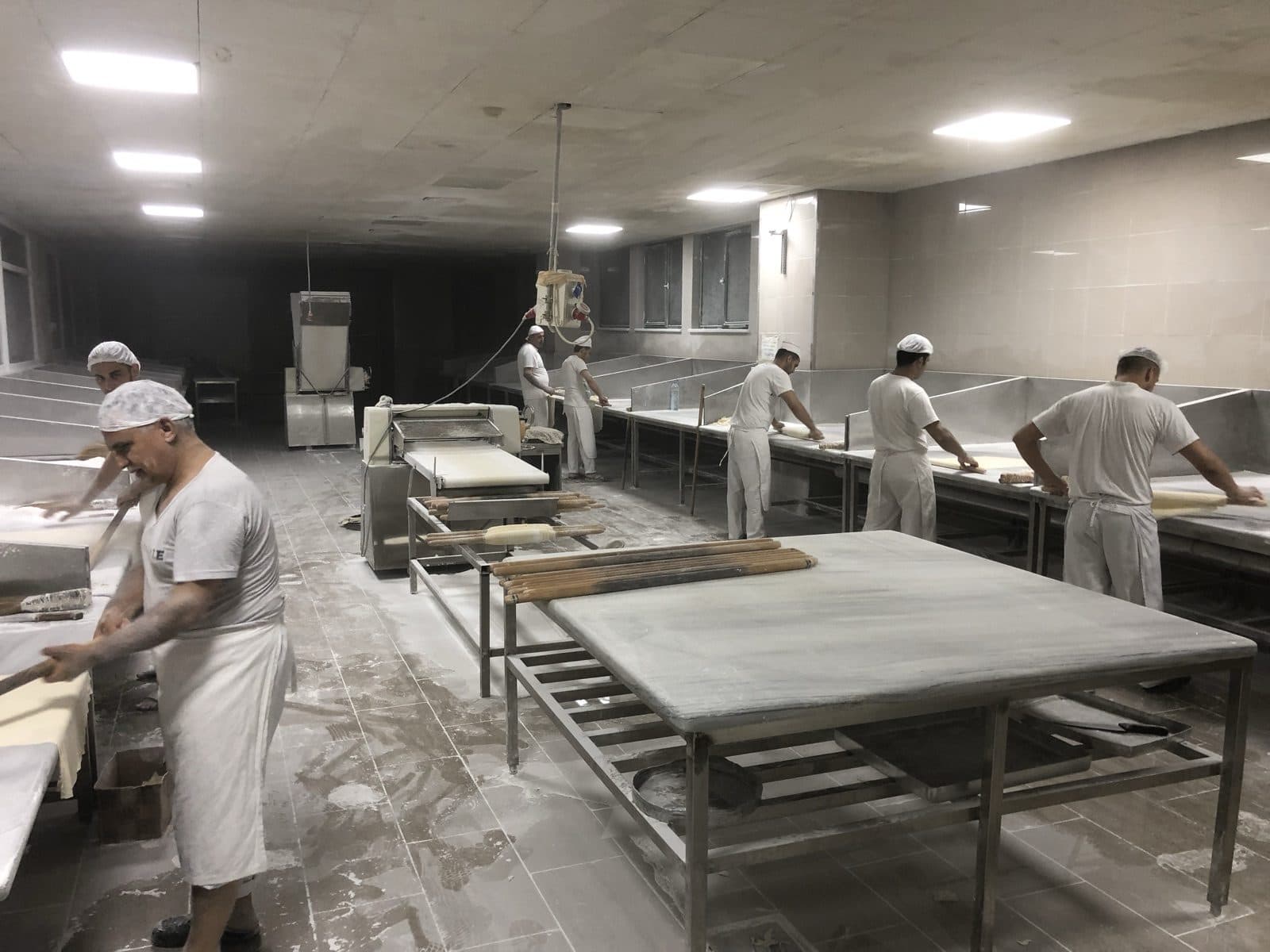 The bakery floor before dawn — bakers in whites working phyllo across long marble tables dusted with flour