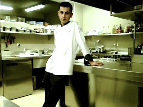 A young Sinan Camozu in chef's whites, leaning against the stainless pass in the hotel kitchen